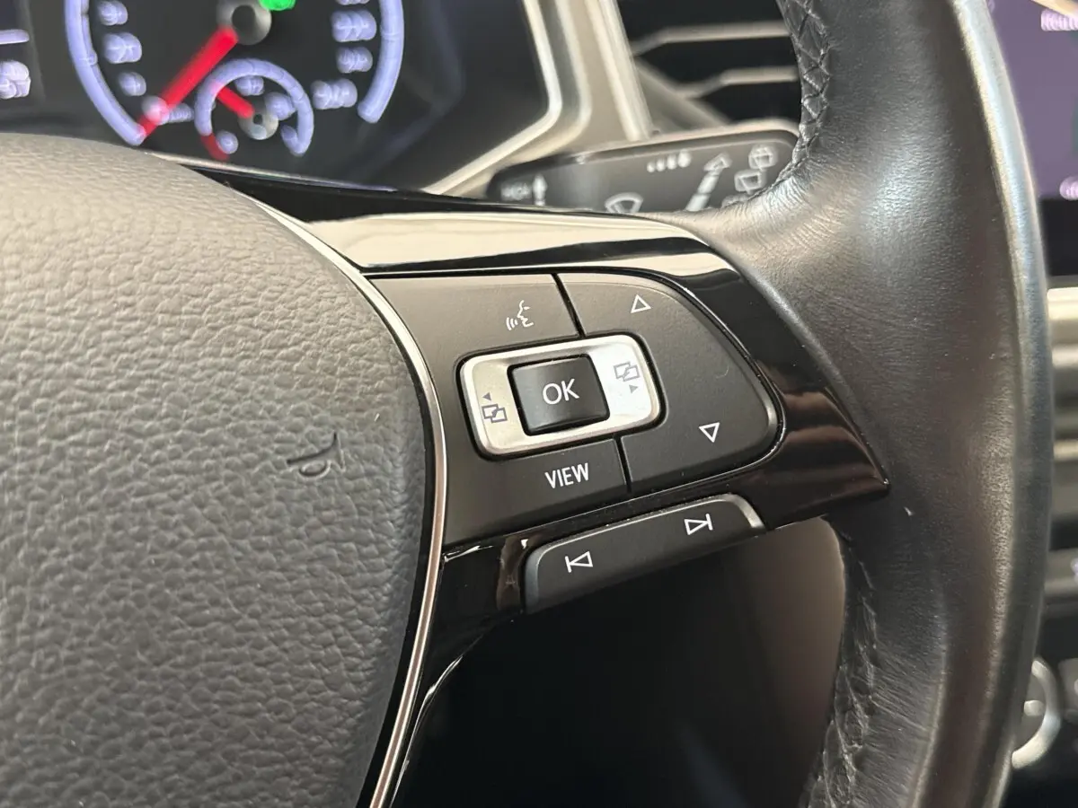Gros plan sur les commandes multifonctions noires et argent du volant cuir du Volkswagen T-Roc 2.0 TDI bleu, tableau de bord en arrière-plan.