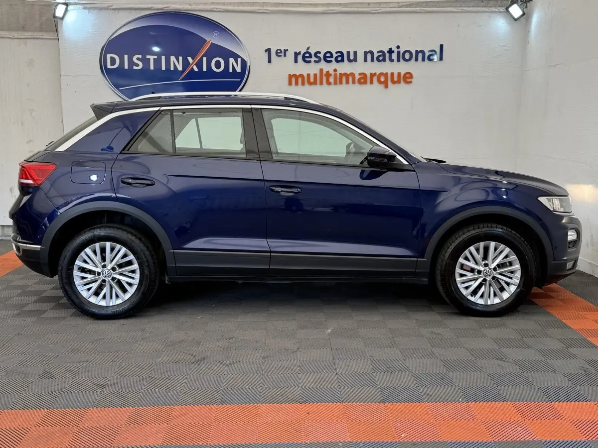 Vue de profil côté gauche d'un Volkswagen T-Roc bleu 2020 stationné en intérieur avec jantes argentées et toit noir.