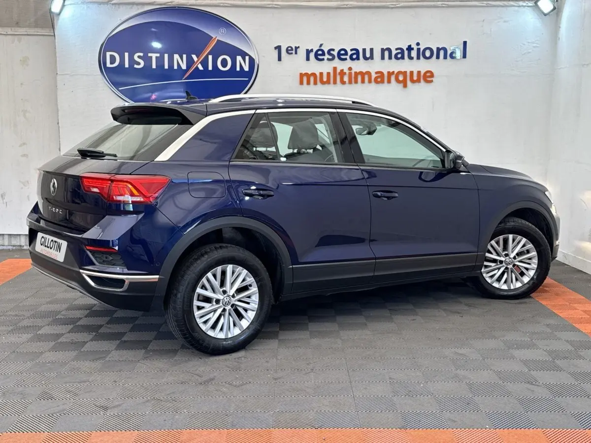 Volkswagen T-Roc bleu en vue profil droit, avec toit noir et jantes alliage, stationné en intérieur showroom.