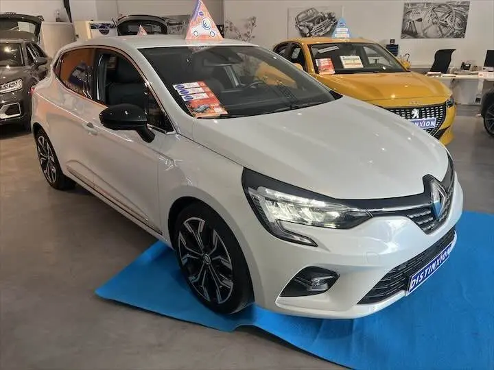 Renault Clio V blanc nacré vue 3/4 avant droit en showroom avec jantes noires et détails noirs contrastants.