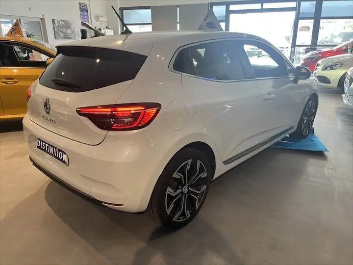 Renault Clio V blanc nacré vue 3/4 arrière droit en showroom avec jantes noires et feux allumés.