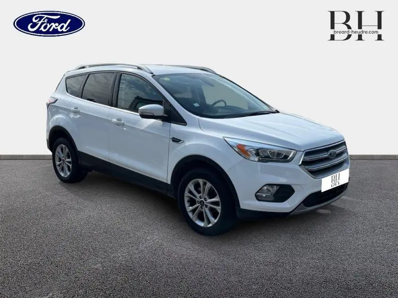 Ford Kuga blanc glacier en 3/4 avant droit, avec barres de toit aluminium et jantes alliage visibles.