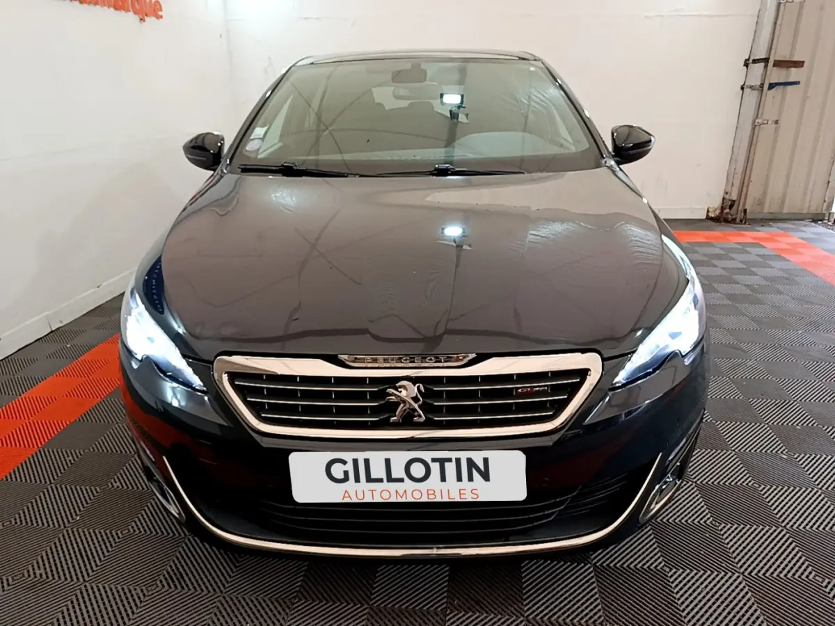 Vue frontale d'une Peugeot 308 gris foncé avec phares LED allumés dans un showroom intérieur.