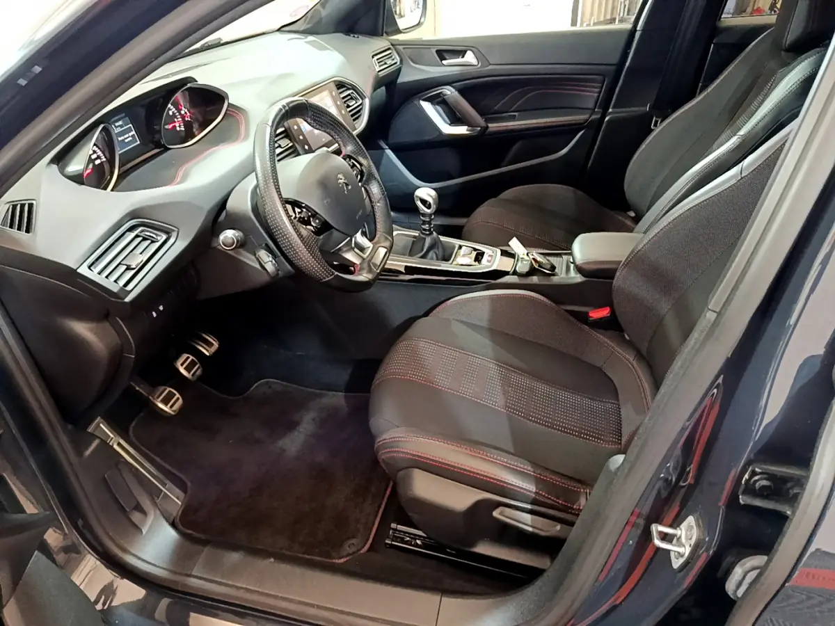 Intérieur avant droit de la Peugeot 308 gris foncé 2016, volant cuir, sièges tissu avec surpiqûres rouges et boîte manuelle.