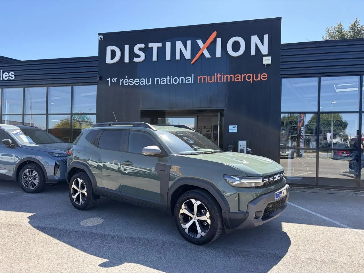 Vue 3/4 avant droite d'un Dacia Duster III hybride 2026 en kaki lichen avec jantes alliage diamantées devant un showroom Distinxion.