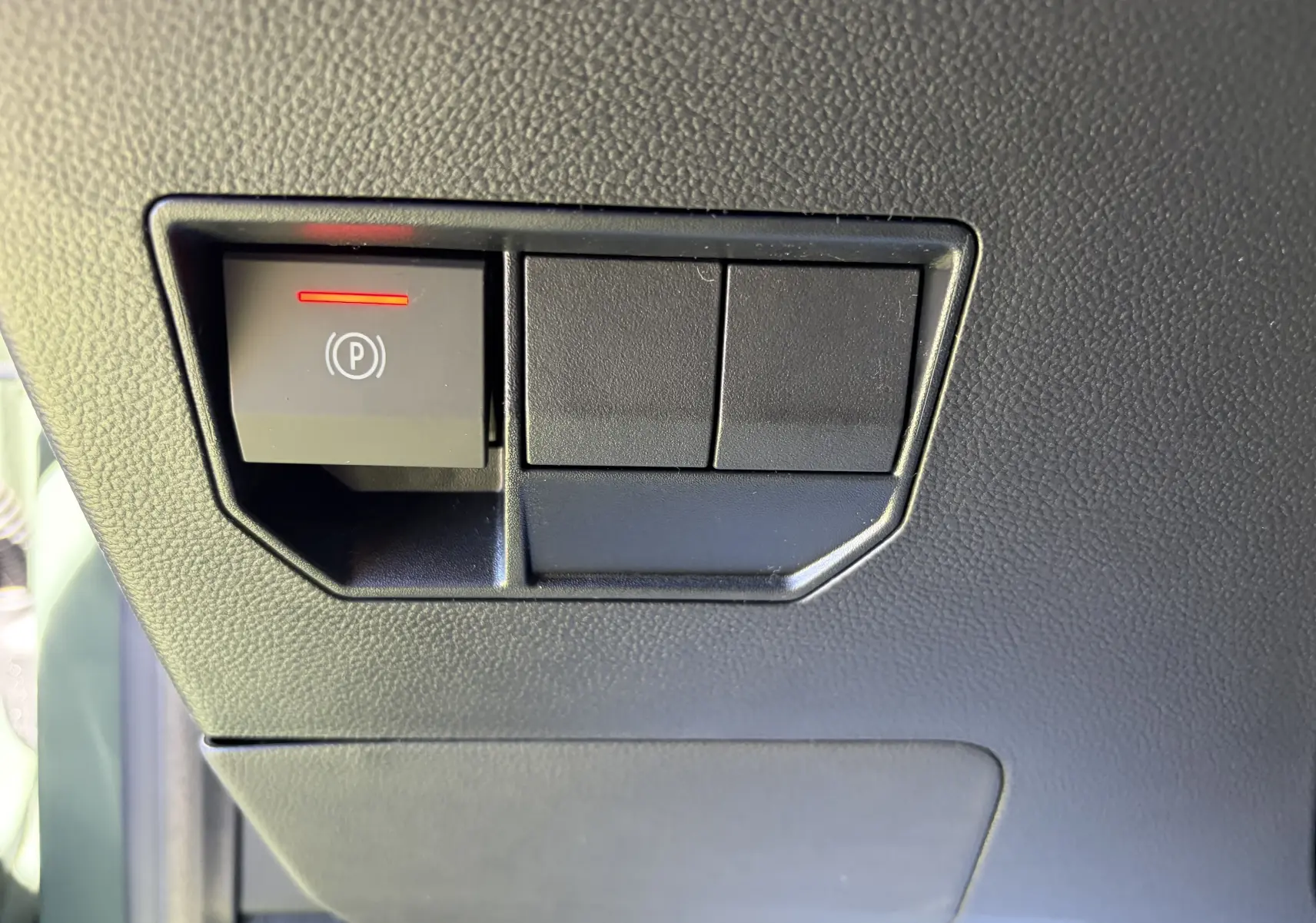 Gros plan sur le bouton de frein de stationnement électrique avec témoin rouge, tableau de bord noir du Dacia Duster III.