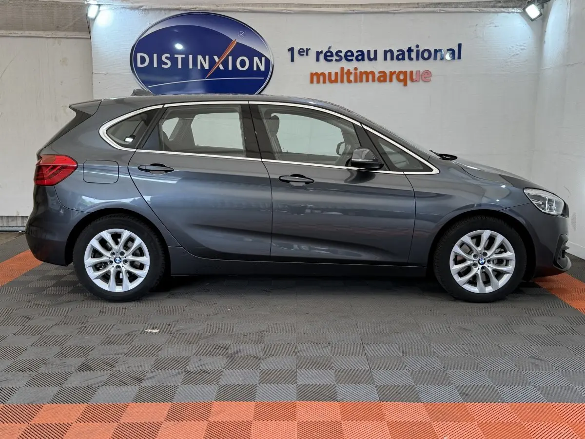 BMW Série 2 Active Tourer gris foncé vue de profil côté gauche, avec jantes alliage et logo Distinxion en arrière-plan.