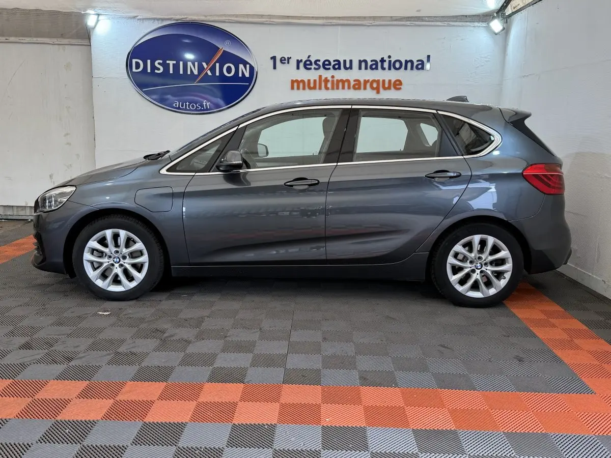 Profil côté gauche d'une BMW Série 2 Active Tourer 225xe gris foncé dans un showroom avec logo Distinxion en arrière-plan.