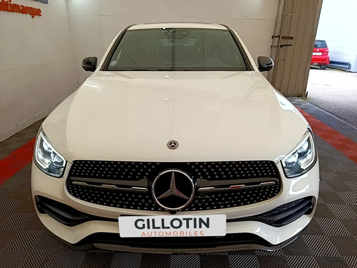 Vue avant d'un Mercedes GLC Coupé blanc avec calandre AMG noire et phares LED allumés dans un garage.