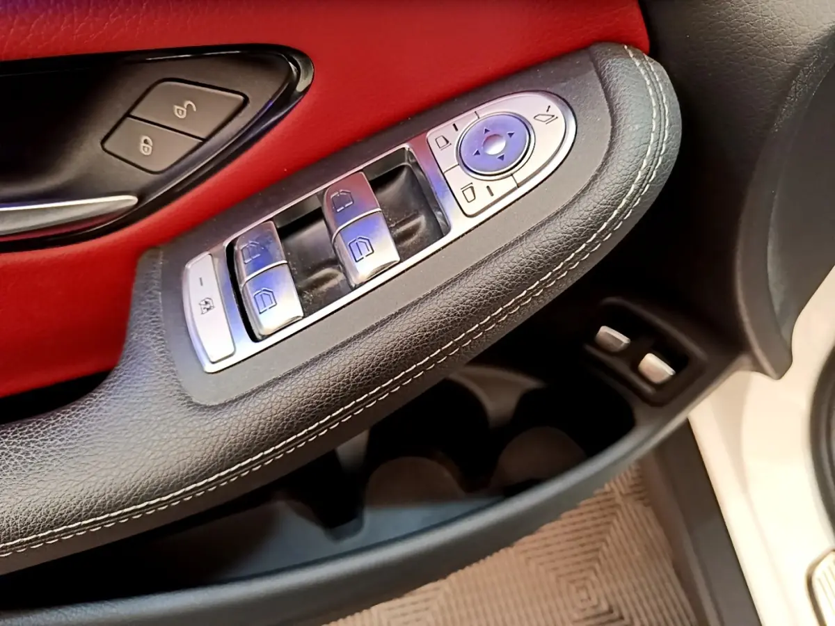 Gros plan sur la commande des vitres et rétroviseurs côté conducteur, garniture cuir noir et panneau rouge, porte blanche visible.
