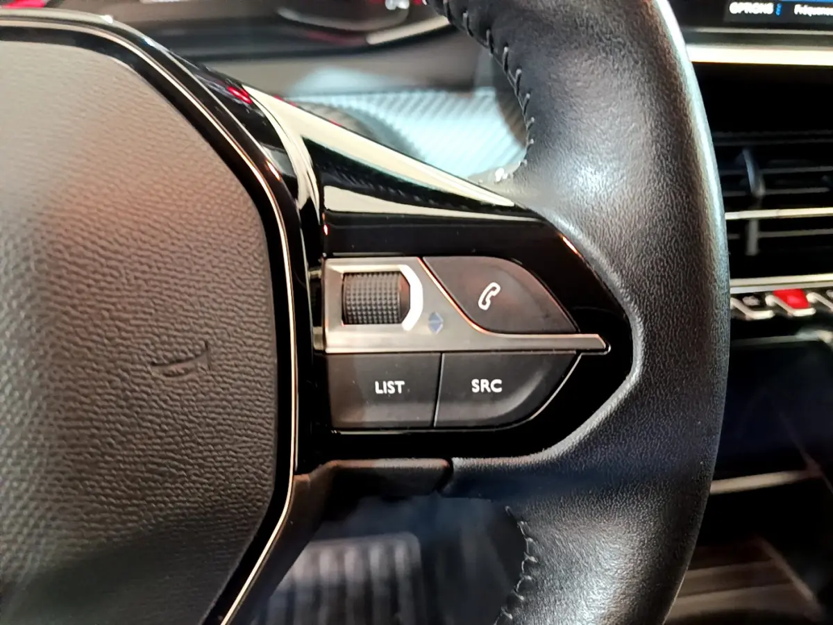 Gros plan sur les commandes gauche du volant cuir noir du Peugeot 2008 gris clair avec tableau de bord en arrière-plan.