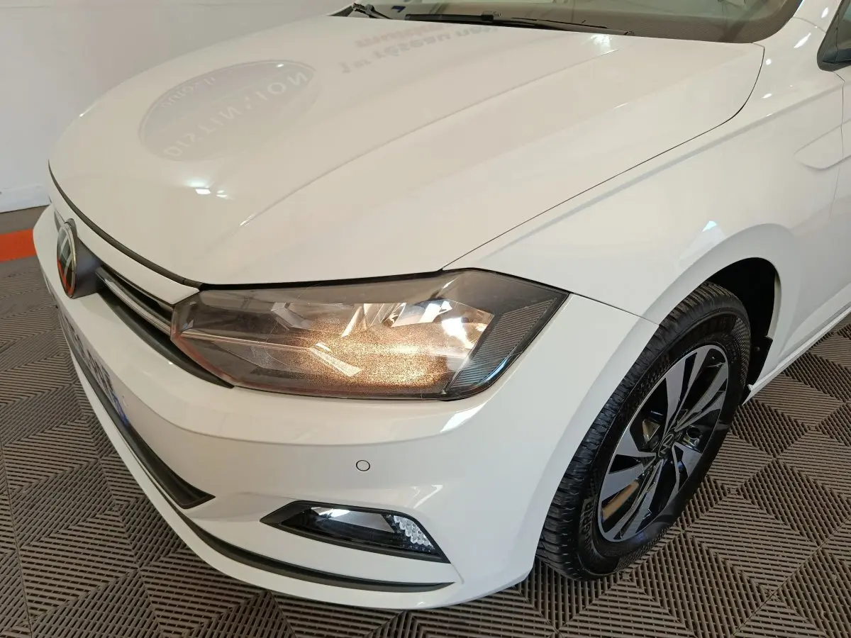 Vue 3/4 avant droit d'une Volkswagen Polo blanche avec phares allumés et jantes noires à motifs modernes.