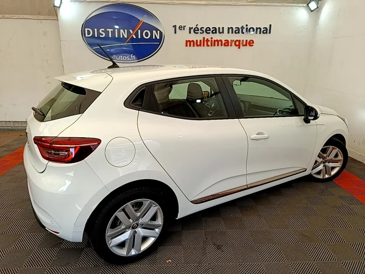 Vue 3/4 arrière droite d'une Renault Clio Business Blue dCi 85 blanche avec jantes alliage et logo Distinxion en arrière-plan.
