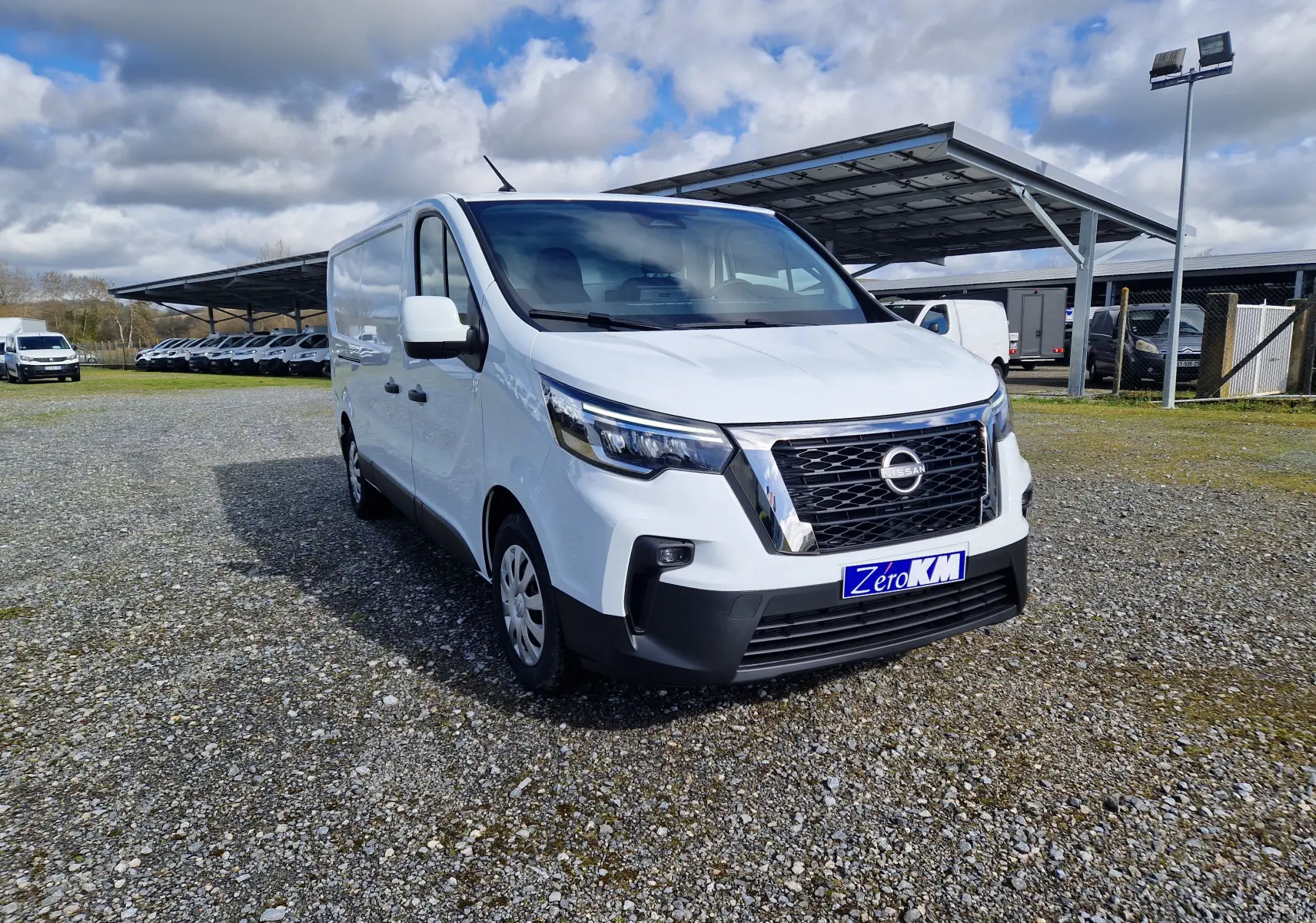 Nissan Primastar blanc en vue 3/4 avant droit sur parking extérieur avec abri métallique en arrière-plan.