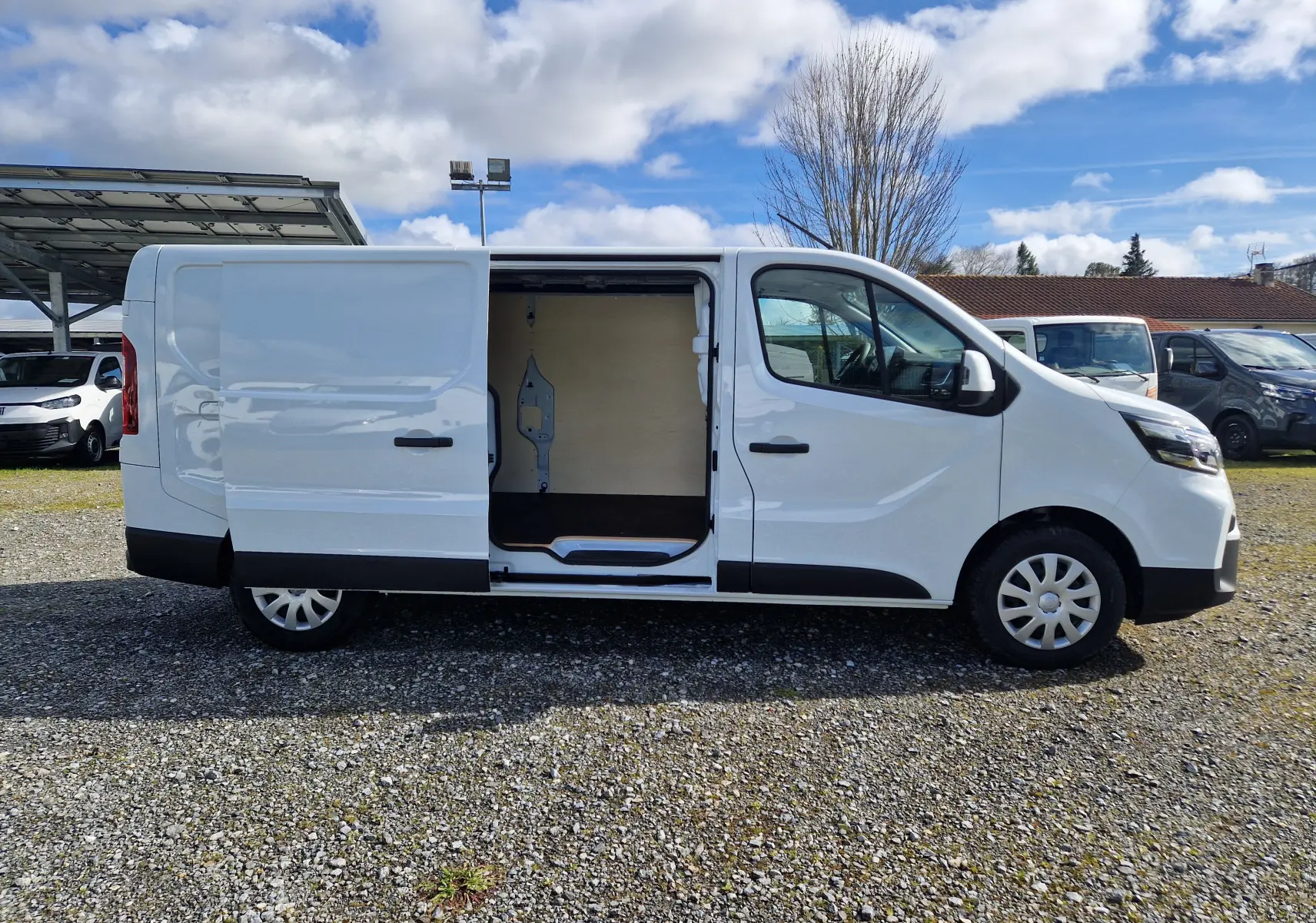 Vue de profil côté gauche d’un utilitaire Nissan Primastar blanc, version L2H1 avec porte latérale coulissante, sur un sol gravillonné.