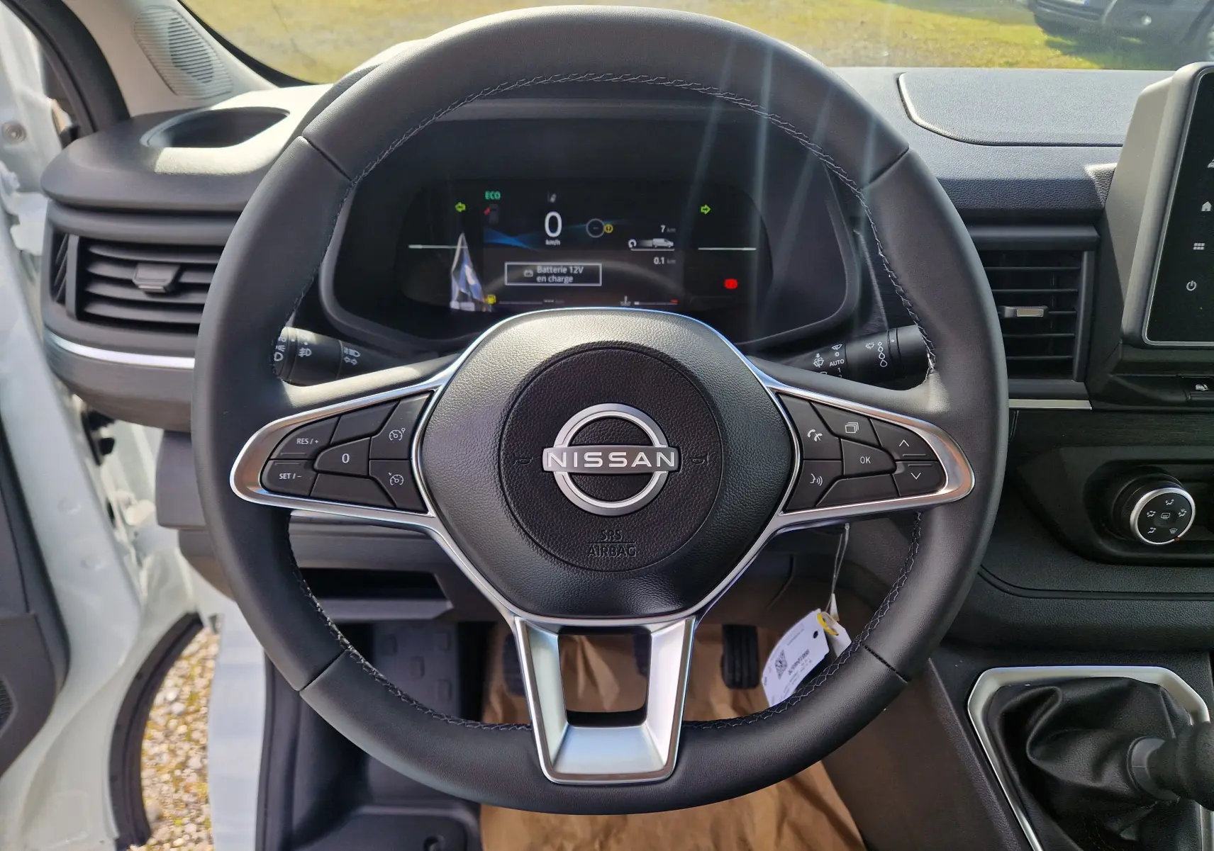 Vue intérieure centrée sur le volant noir du Nissan Primastar blanc, avec tableau de bord digital et levier de vitesses manuel visible.