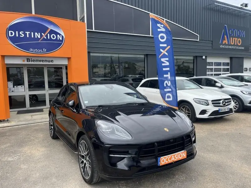 Porsche Macan noir vue 3/4 avant droit sur parking devant concession avec drapeau Distinxion.