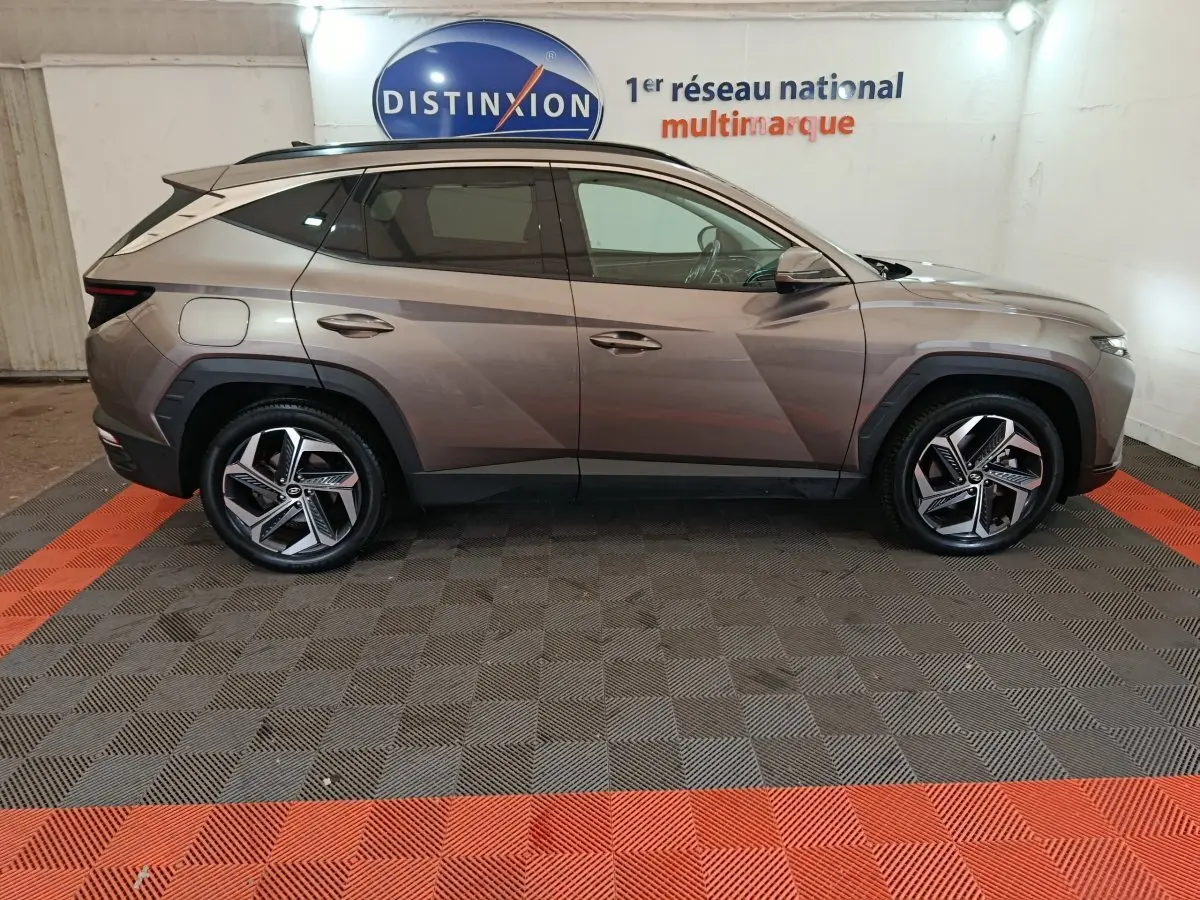 Vue latérale côté gauche d'un Hyundai Tucson marron 2022 hybride rechargeable sur sol damier gris et rouge en intérieur showroom.
