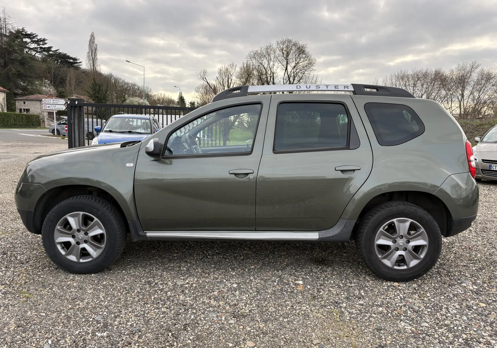 Vue de profil côté gauche d'un Dacia Duster 1.5 DCI 110 vert foncé avec barres de toit et jantes noires sur un sol gravillonné.