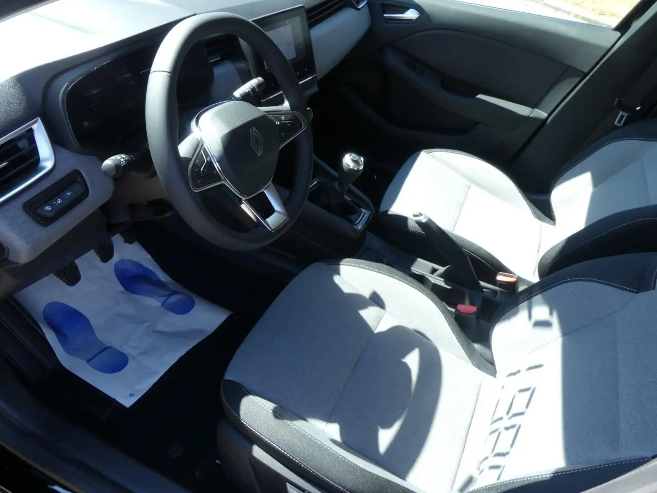 Intérieur avant droit de Renault Clio 2025, sièges gris, volant multifonction, levier de vitesse manuel et écran tactile visible.