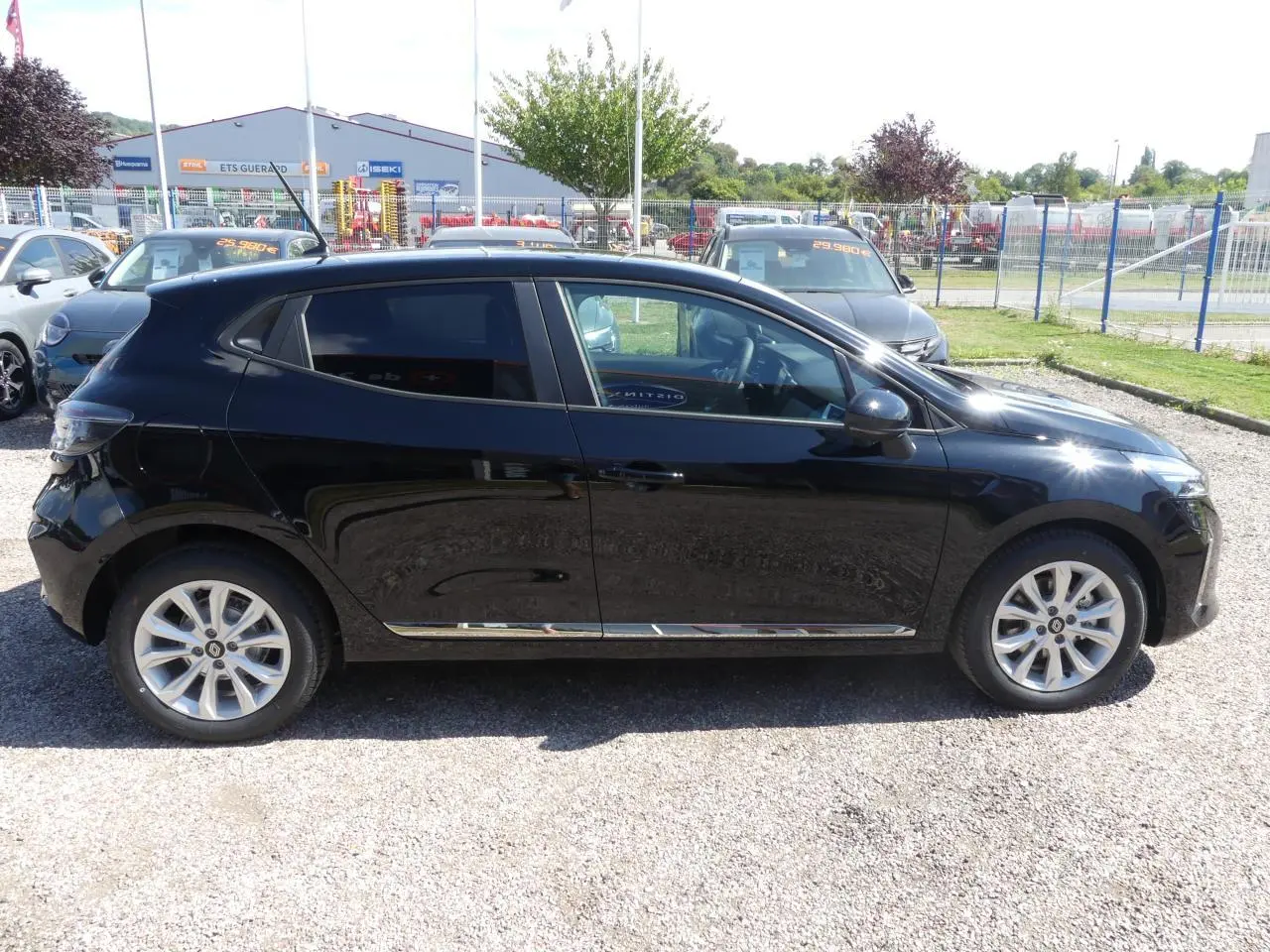 Profil côté gauche d'une Renault Clio noire métallisée 2025 garée sur un sol gravillonné en extérieur.