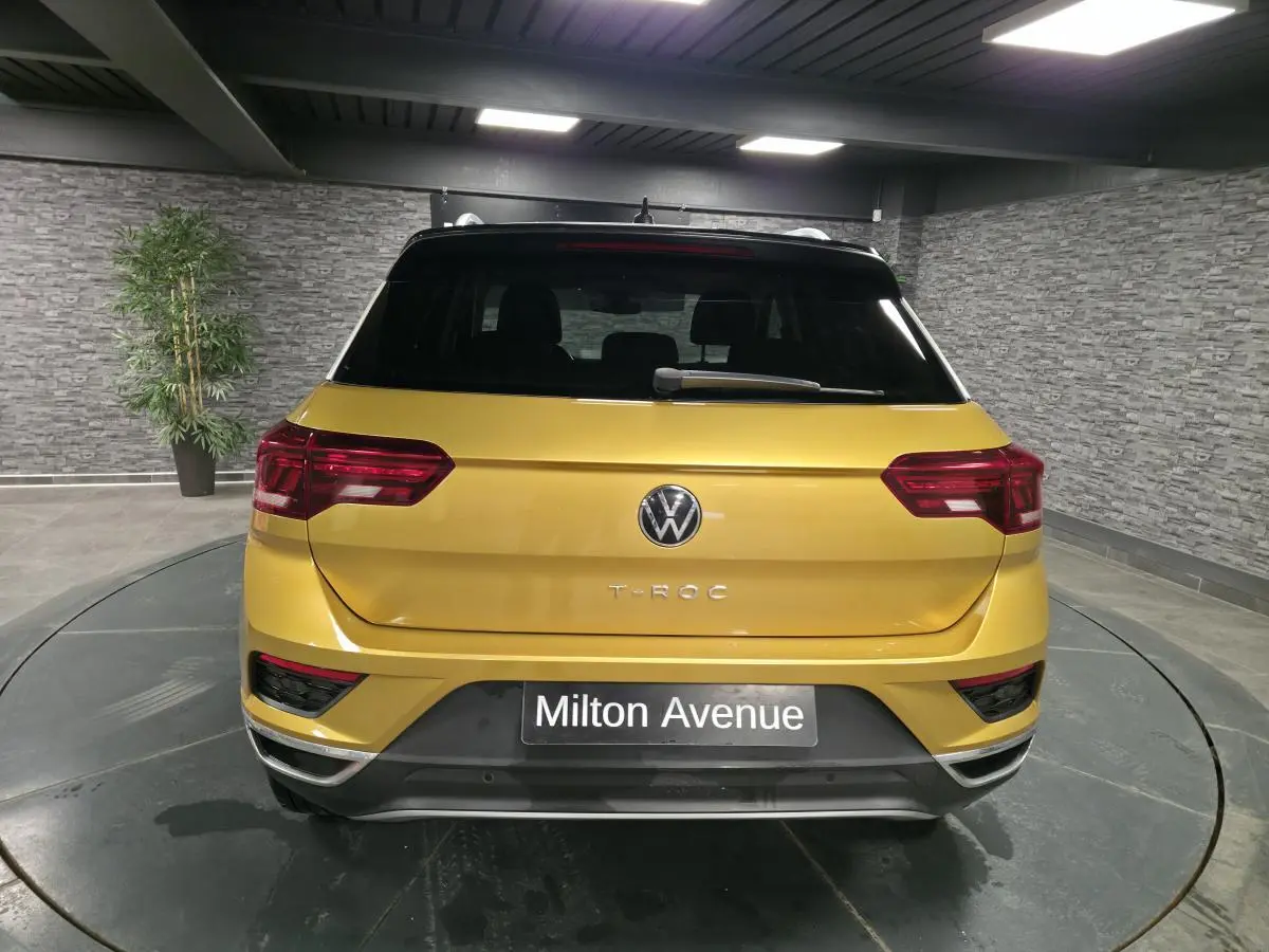 Vue arrière d'un Volkswagen T-Roc jaune Curcuma avec toit noir et feux arrière LED dans un showroom.