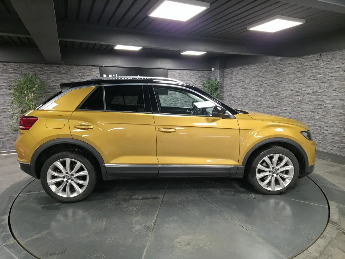 Profil côté gauche d'un Volkswagen T-Roc jaune Curcuma avec toit noir et jantes alliage 18 pouces en intérieur showroom.
