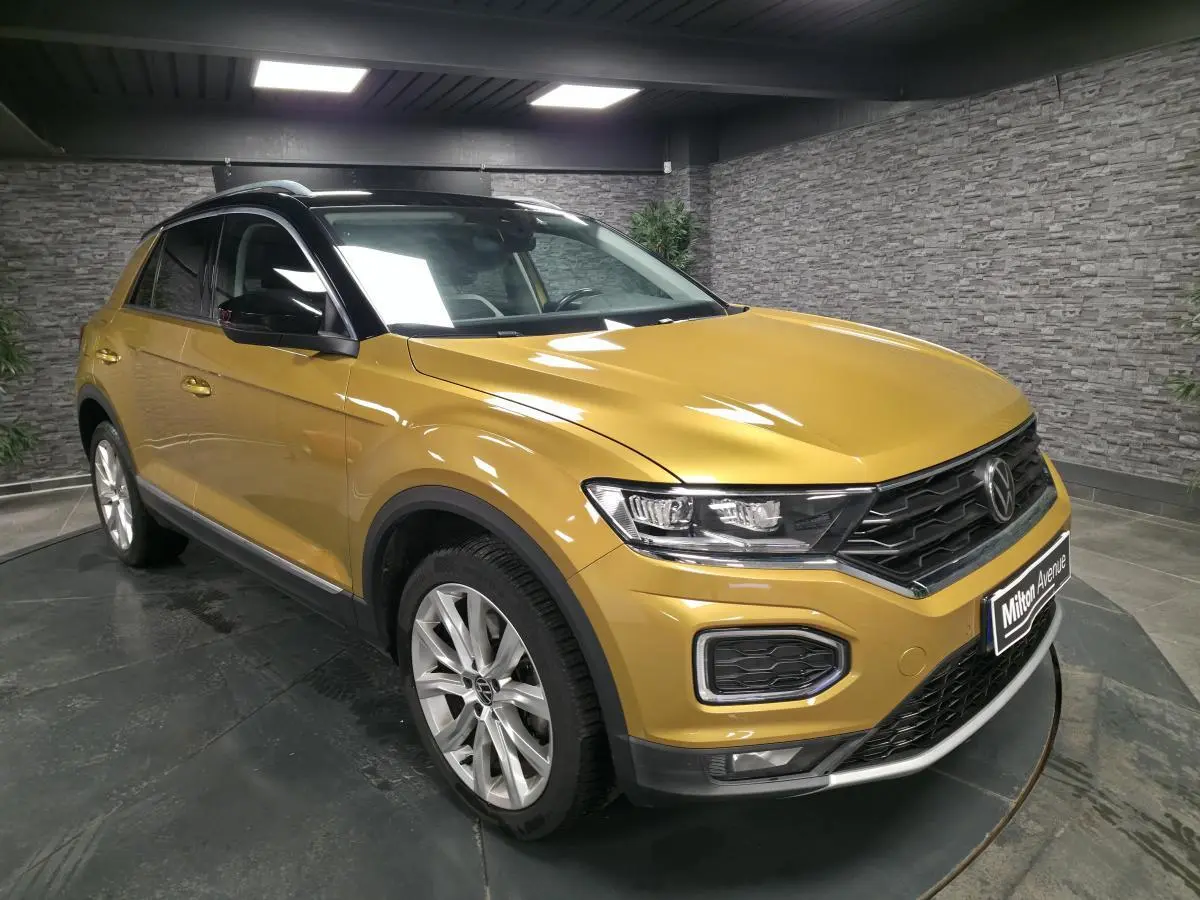 Volkswagen T-Roc jaune Curcuma en 3/4 avant droit avec toit noir et jantes alliage 18 pouces dans un showroom.