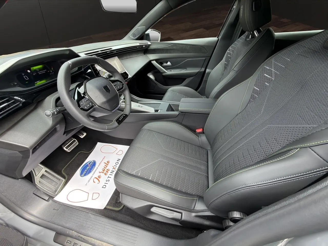 Vue intérieure côté conducteur de la Peugeot 308 GT 2026, sièges noirs avec surpiqûres vertes et tableau de bord numérique.