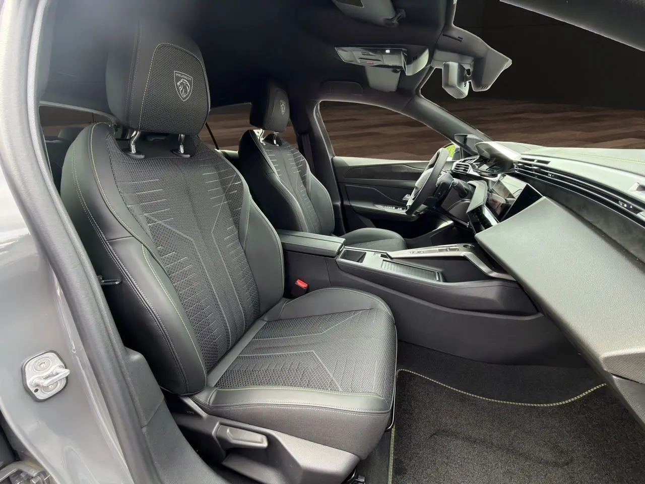 Vue intérieure côté conducteur de la Peugeot 308 GT 2026 gris selenium, sièges noirs avec surpiqûres vertes et tableau de bord moderne.