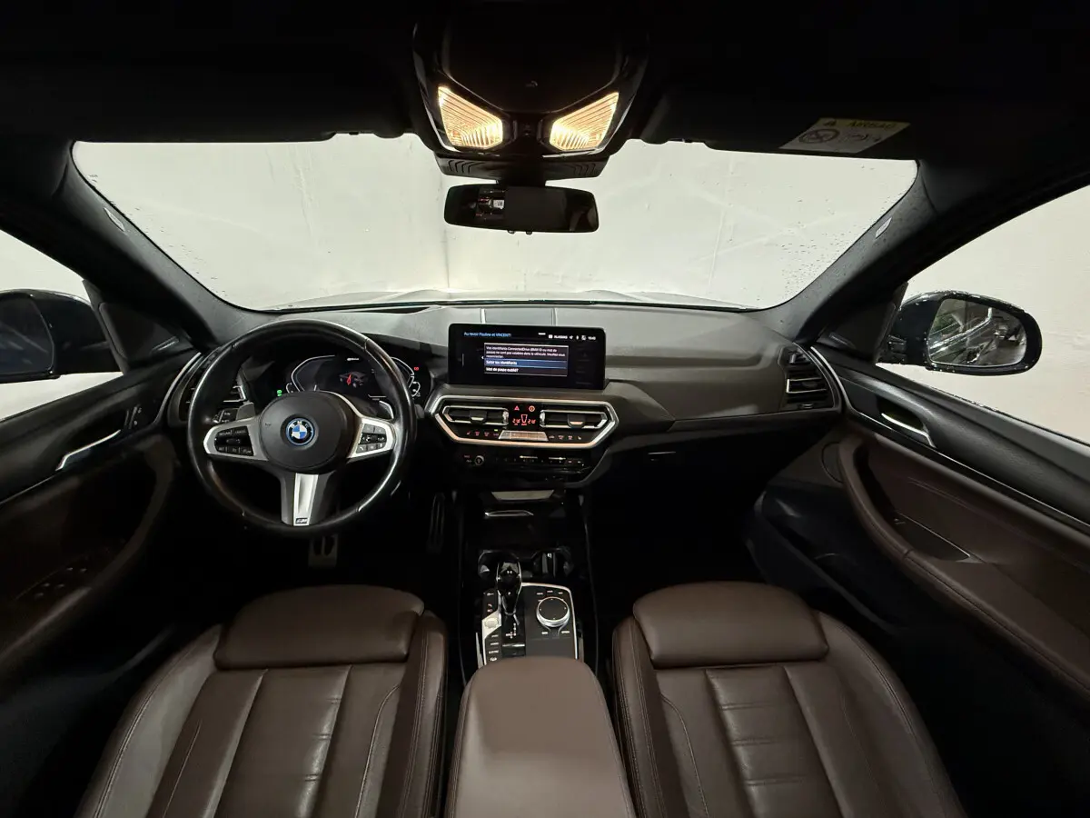 Intérieur BMW X3 xDrive30e 2023 vu de face, tableau de bord noir avec sièges cuir marron et écran tactile central.