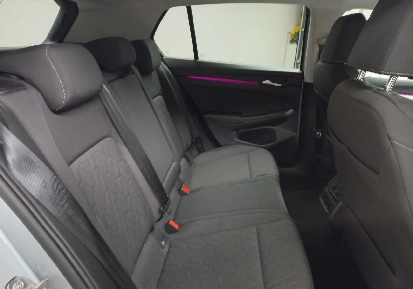 Vue intérieure arrière de la Volkswagen Golf 1.5 eTSI 2025, sièges tissu gris et éclairage d’ambiance violet sur la porte.