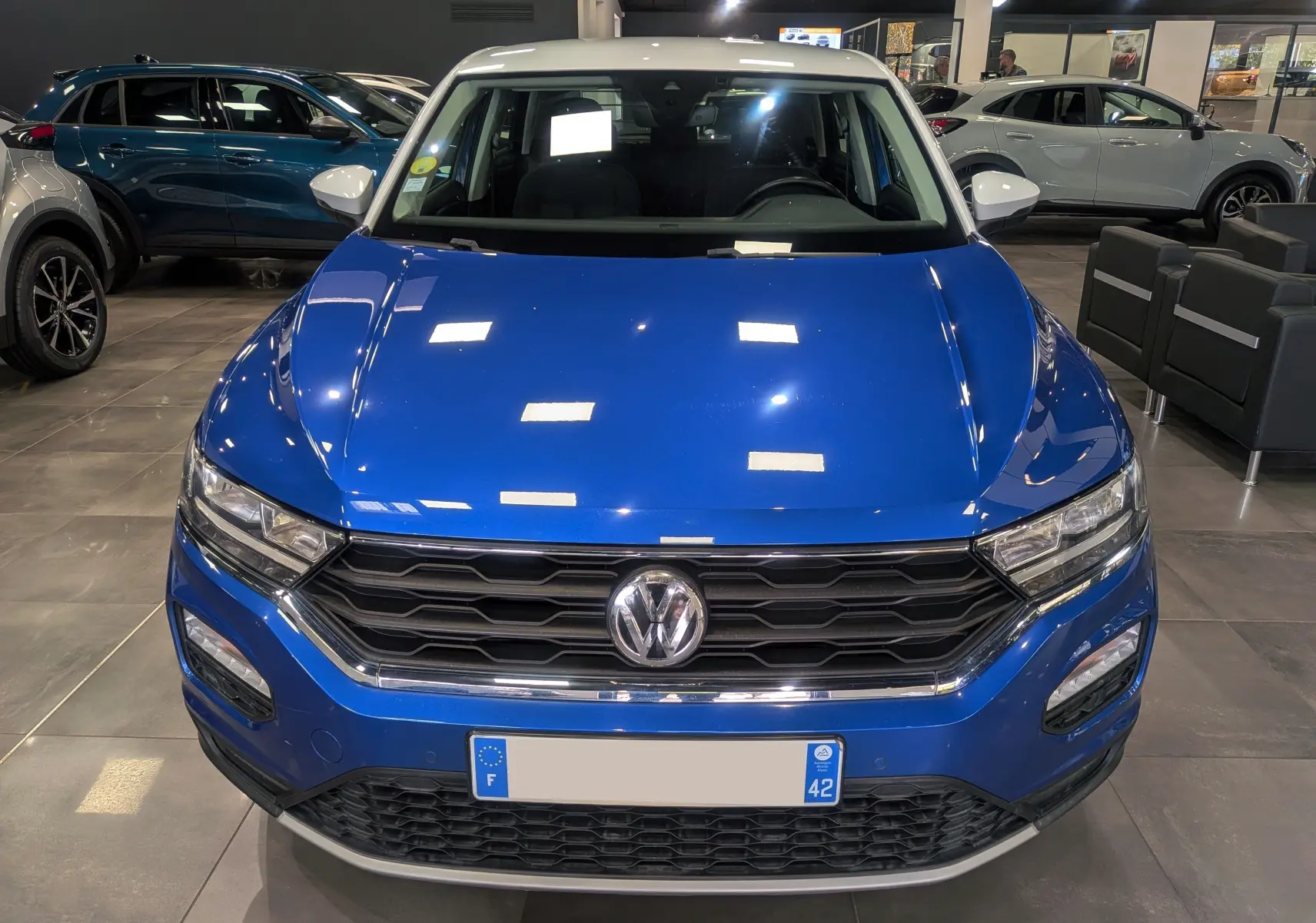 Vue frontale d'un Volkswagen T-Roc bleu Ravenna avec toit blanc, en showroom, mettant en valeur sa calandre hexagonale.