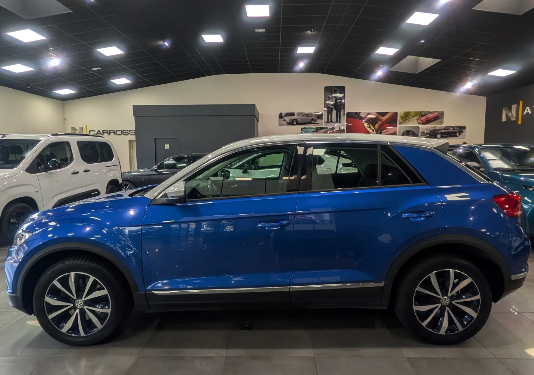 Profil côté gauche d'un Volkswagen T-Roc bleu Ravenna avec toit blanc, roues alliage et détails chromés en showroom.