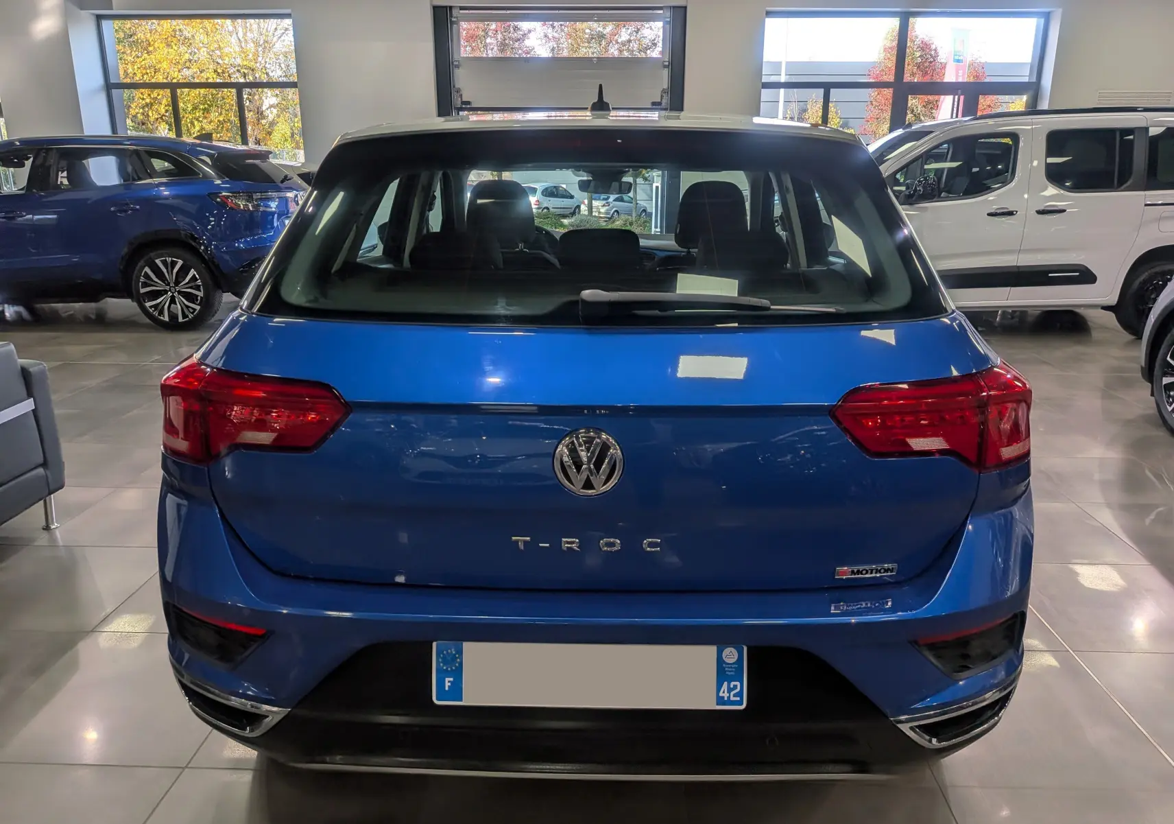 Vue arrière d'un Volkswagen T-Roc bleu Ravenna avec toit blanc, badges T-Roc et 4MOTION visibles en intérieur showroom.
