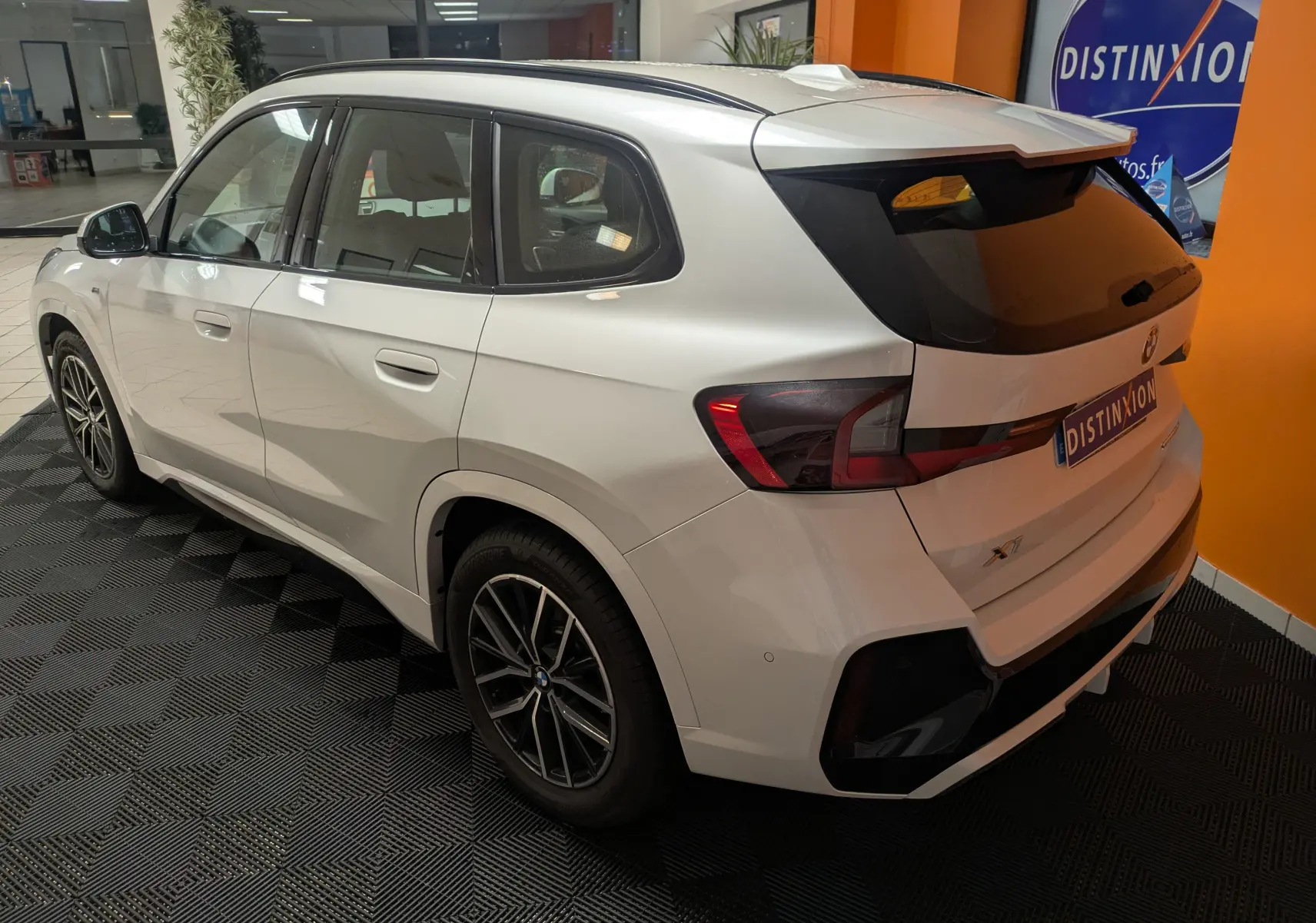 Vue 3/4 arrière droite d’un BMW X1 blanc 2025 avec jantes bicolores et vitres arrière teintées en intérieur showroom.