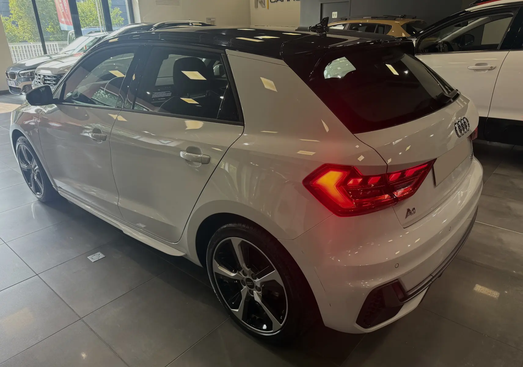 Audi A1 30 TFSI S-Line blanche avec toit noir, vue 3/4 arrière droit en intérieur showroom, feux arrière allumés.