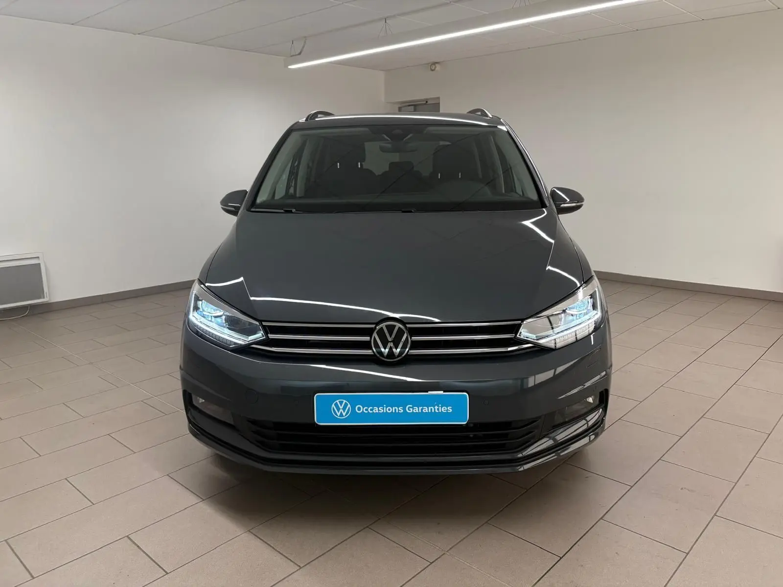 Vue frontale d'un Volkswagen Touran gris dauphin 2025 avec phares LED allumés dans un intérieur neutre.