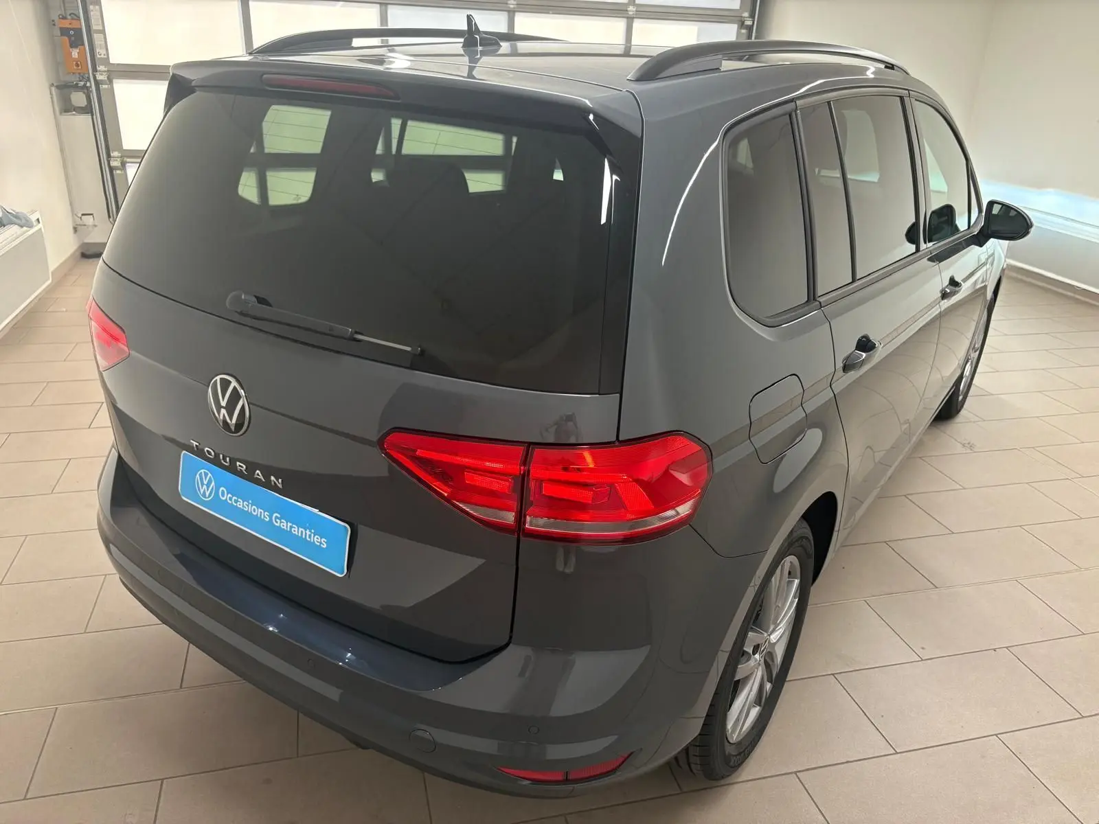 Vue 3/4 arrière droite d'un Volkswagen Touran gris dauphin 2025 avec feux LED et hayon électrique en intérieur showroom.