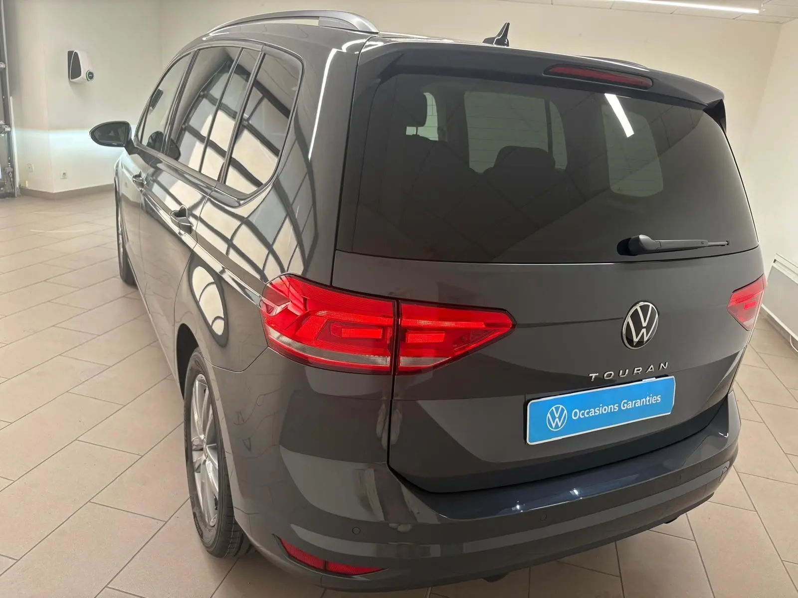 Vue 3/4 arrière gauche d'un Volkswagen Touran gris dauphin avec feux LED et hayon fermé en intérieur showroom.