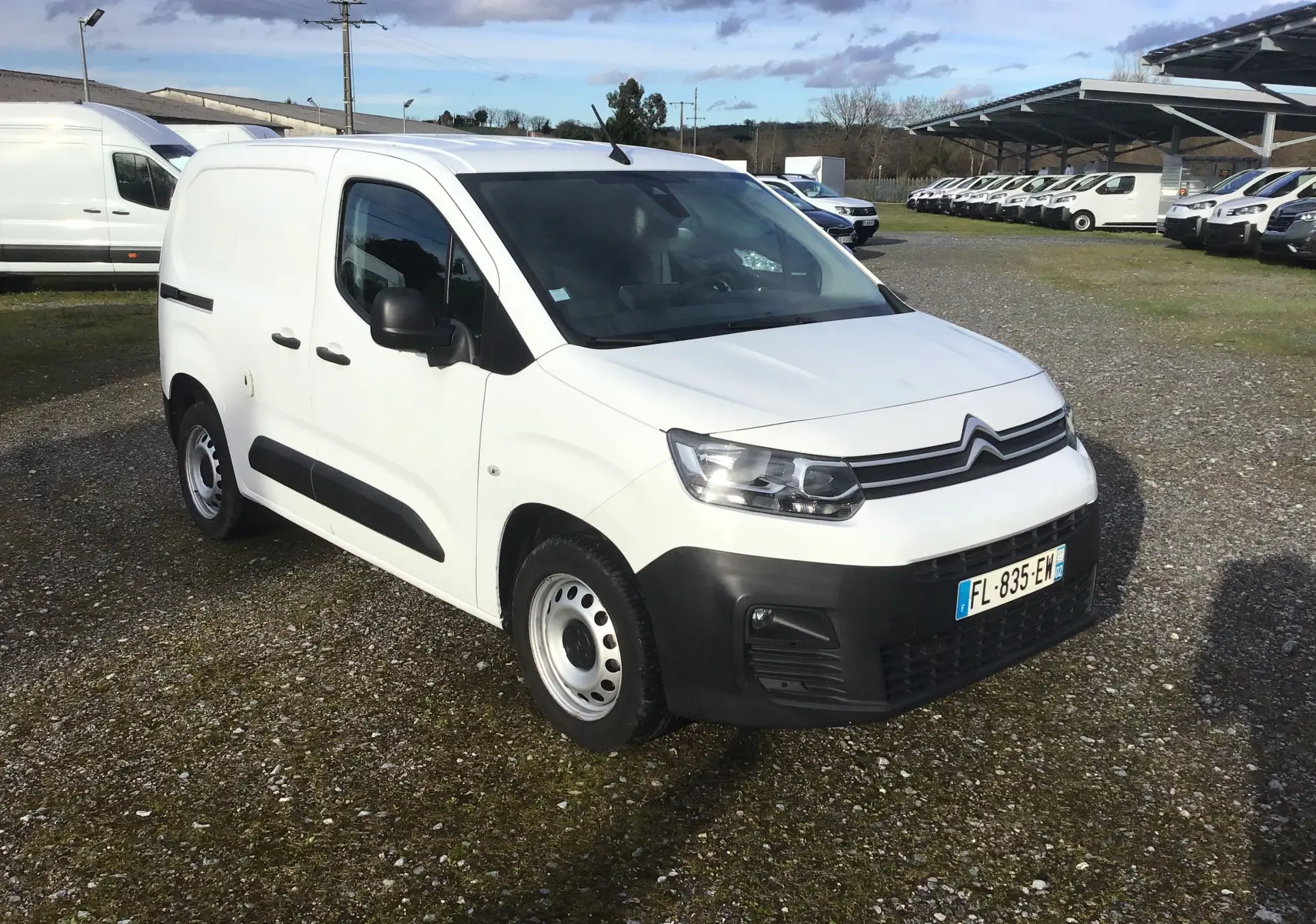 Vue 3/4 avant d’un utilitaire Citroën Berlingo blanc avec pare-chocs noirs, stationné sur un sol gravillonné.