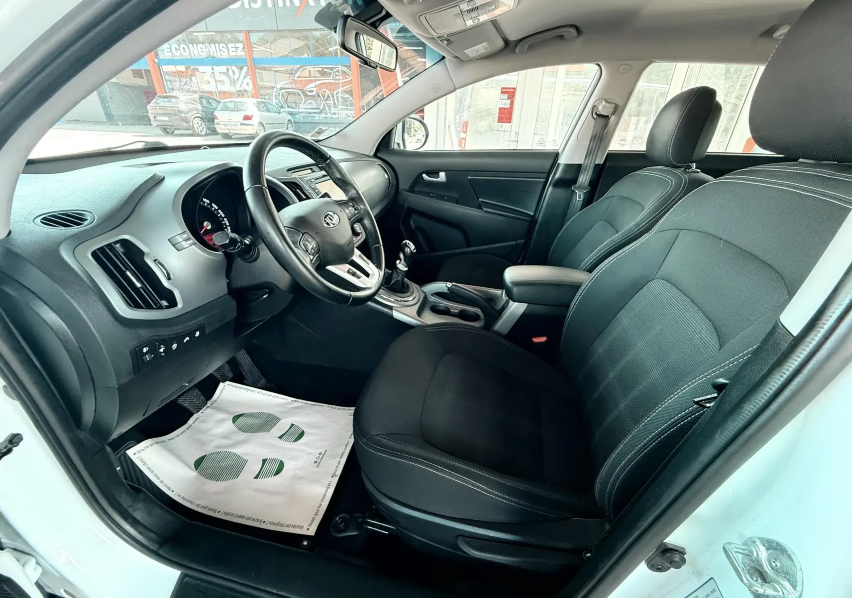 Vue intérieure côté conducteur du Kia Sportage blanc 2015, sièges tissu noir et tableau de bord moderne.