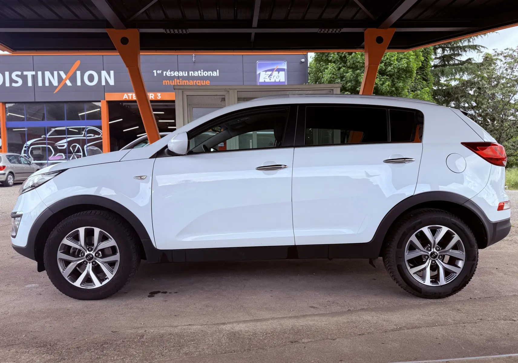 Profil latéral droit d'un KIA Sportage blanc de 2015, avec jantes alliage et poignées chromées sous abri.