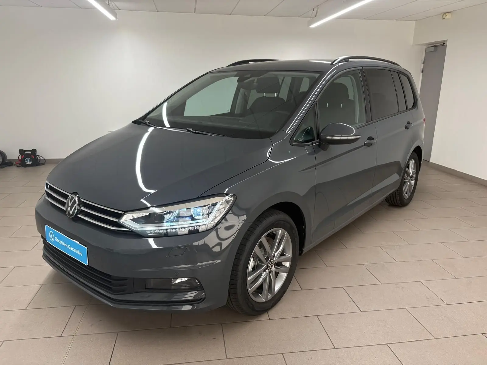 Volkswagen Touran 2025 gris dauphin en vue 3/4 avant droit dans un intérieur lumineux avec jantes alliage 17 pouces.