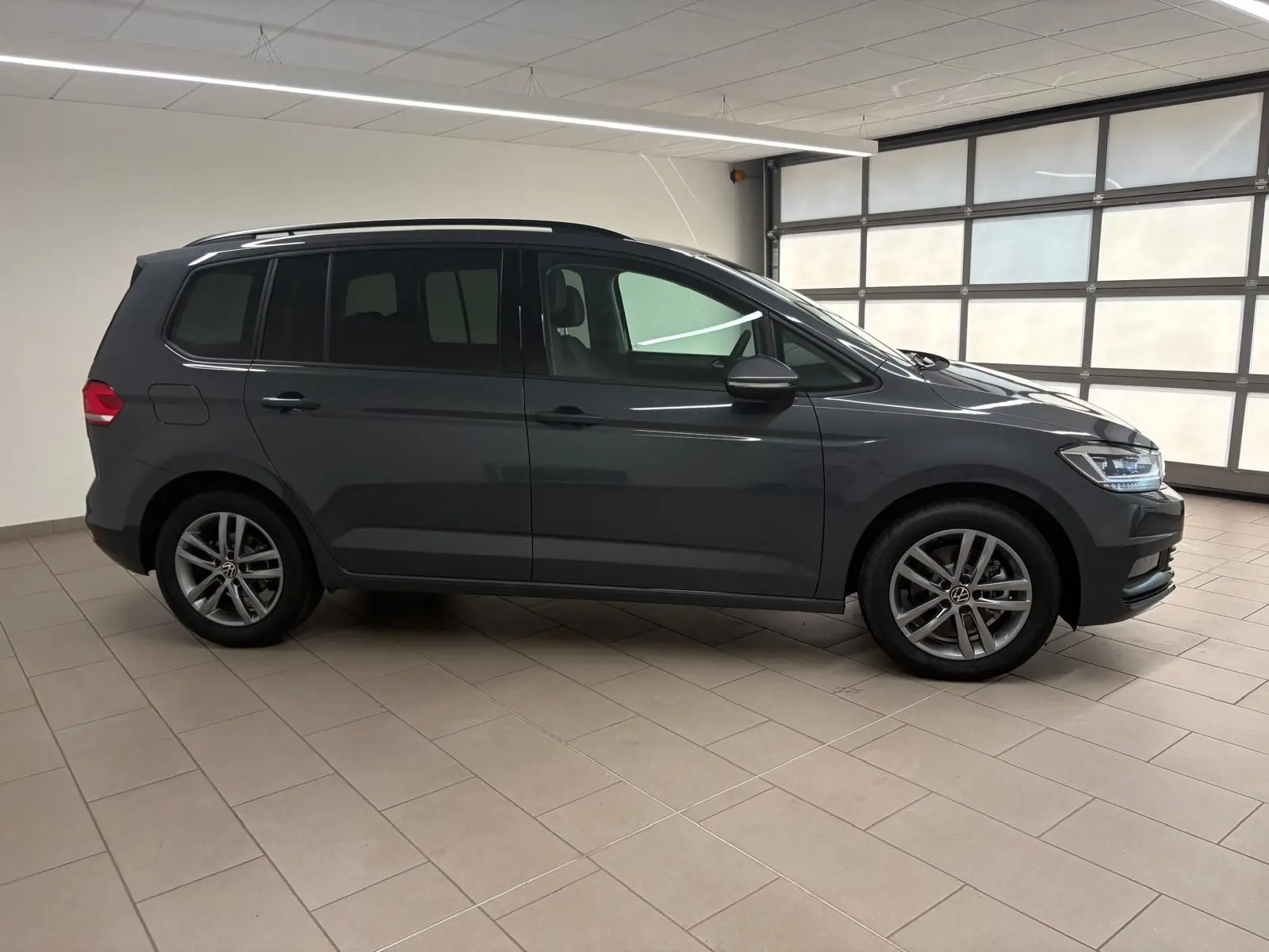 Vue de profil côté gauche d'un Volkswagen Touran gris dauphin 2025 en intérieur, avec jantes alliage 17 pouces et feux LED.
