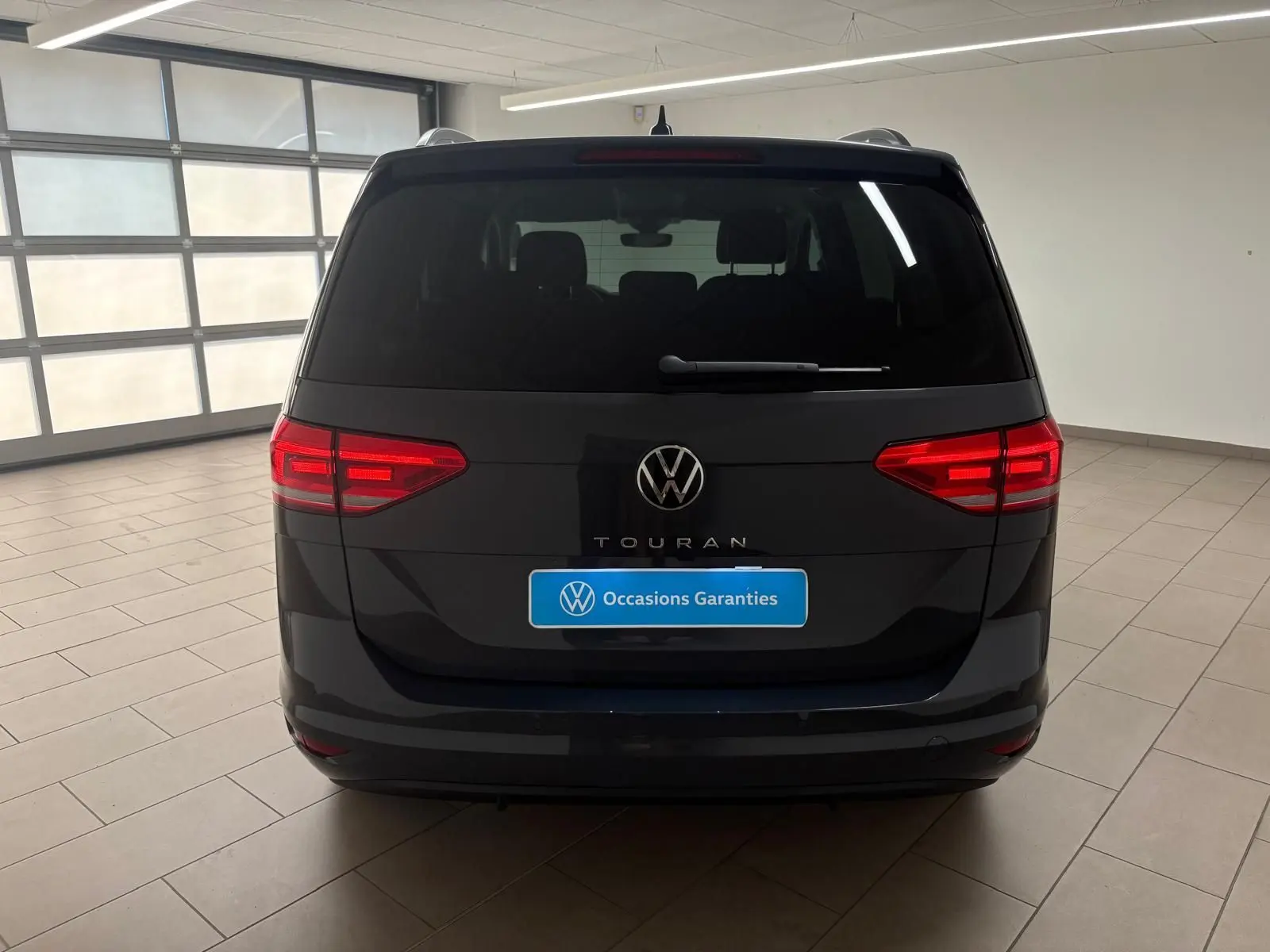 Vue arrière d'un Volkswagen Touran gris dauphin 2025 avec feux arrière allumés en intérieur showroom.