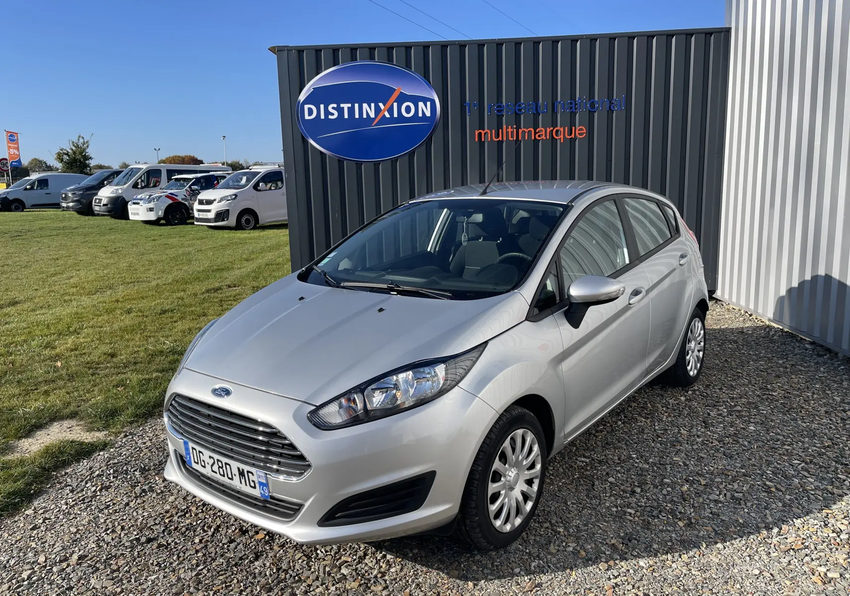 Ford Fiesta 1.25 82 CV Trend gris titane vue 3/4 avant droit sur parking gravillonné devant un mur noir.