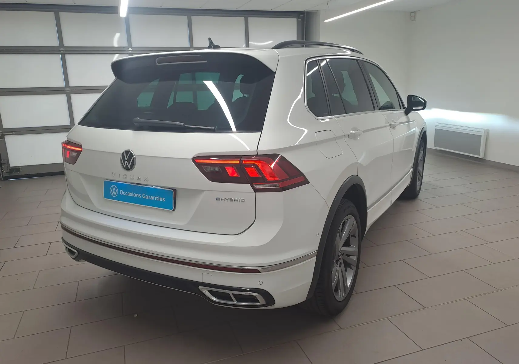 Vue 3/4 arrière droite du Volkswagen Tiguan blanc pur 2023 hybride rechargeable avec finition R-Line en intérieur showroom.