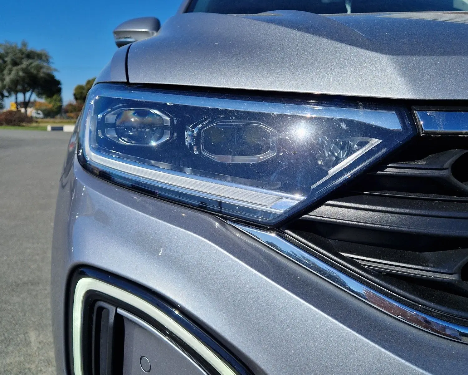 Gros plan sur le phare avant gauche et la calandre d'un Volkswagen T-Roc gris pyrite sous un ciel bleu.