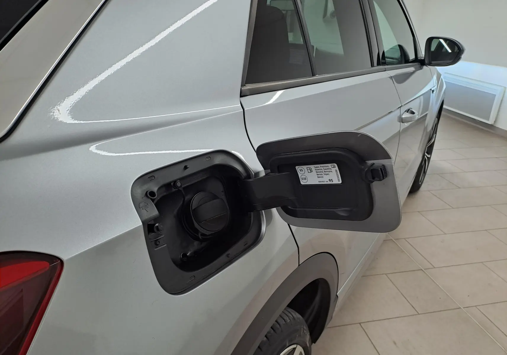Vue arrière côté gauche du Volkswagen T-Roc gris pyrite avec trappe à carburant ouverte sur un sol carrelé intérieur.